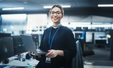 A smiling cybersecurity professional holding a laptop in a high-tech office environment, with multiple monitors displaying analytical data, symbolizing the cutting-edge expertise gained in Herzing University’s cybersecurity program.